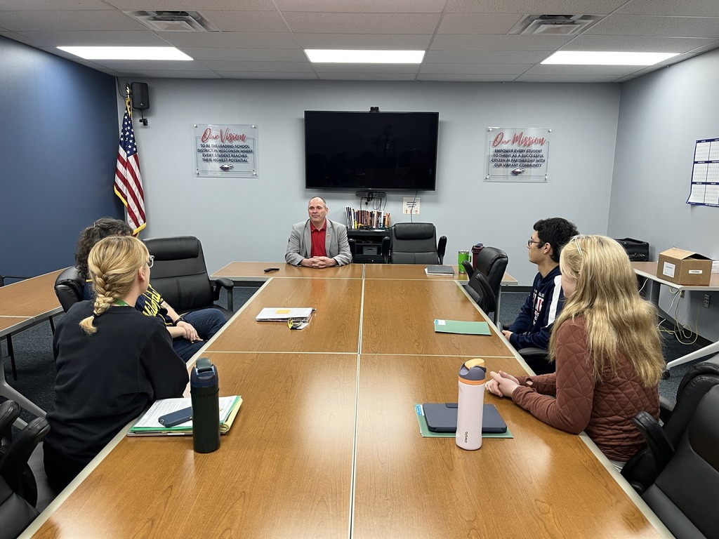 Representative Duke Tucker seated at the head of a conference table, speaking with Unity Raise Your Voice Club members and advisors in a school meeting room.