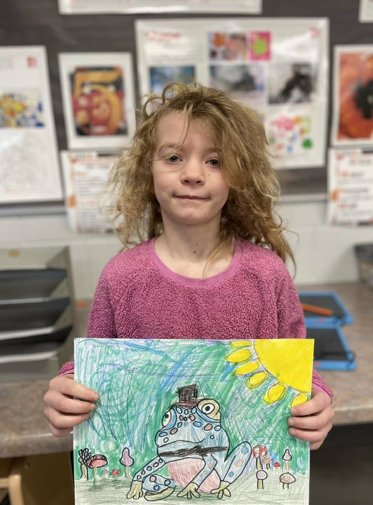 A smiling third-grade student named Marley holding up her colorful drawing of a frog wearing a top hat, featuring a bright yellow sun and a green-textured background.