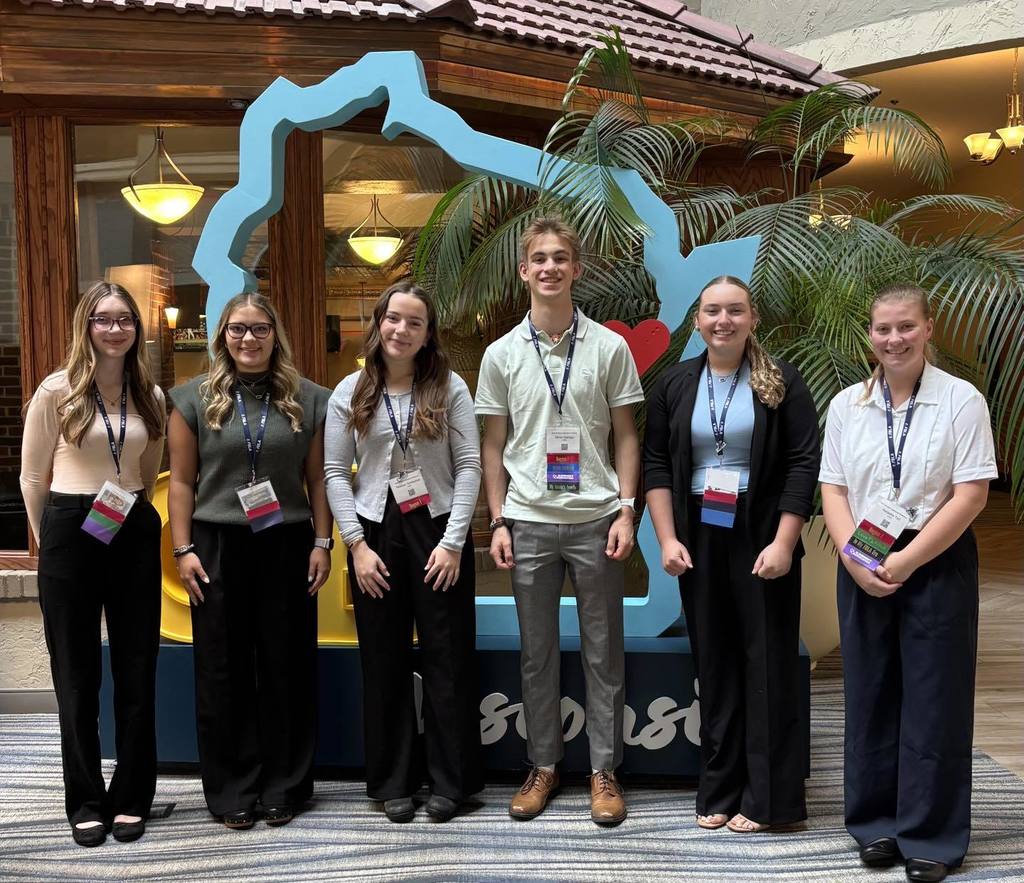 Six students standing and smiling in front of a large blue outline of the state of Wisconsin at the FBLA State conference or event.