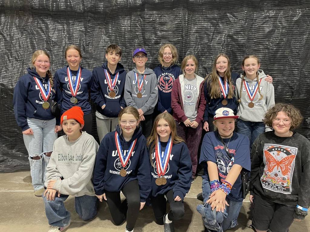 Thirteen middle and high school students from Unity School District posing for a team photo. Several students are wearing gold, silver, or bronze medals over their Science Olympiad hoodies, standing against a black mesh background.