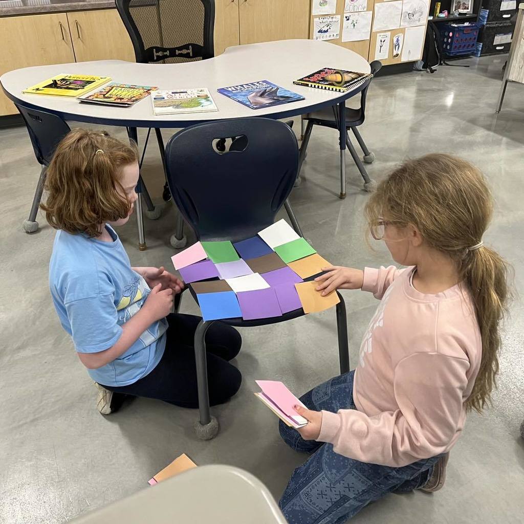 Two students practicing area measurement by tiling the seat of a blue classroom chair with colorful square units.