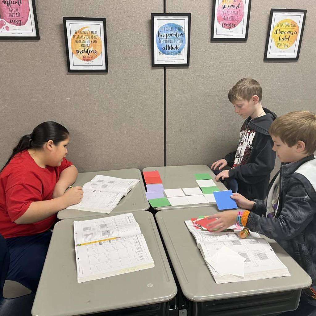Three Unity students collaborating to solve an area problem by tiling their desks with colorful square units. Motivational posters are visible on the wall behind them as they work in a small group.
