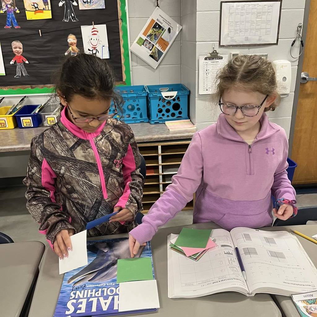 Two Unity students using square tiles to calculate the area of a book cover during a hands-on third-grade math activity.