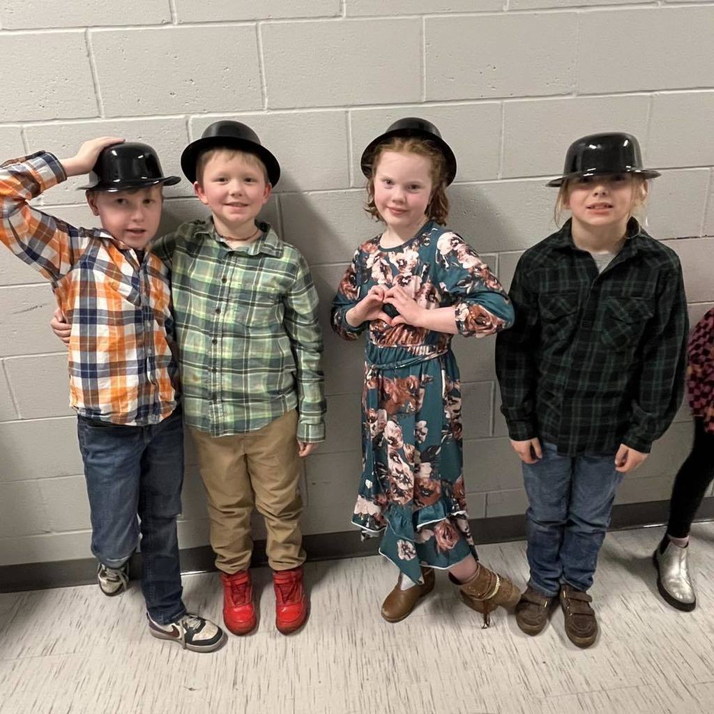 group of four Unity third graders wearing black bowler hats and smiling for a photo against a white cinderblock wall.