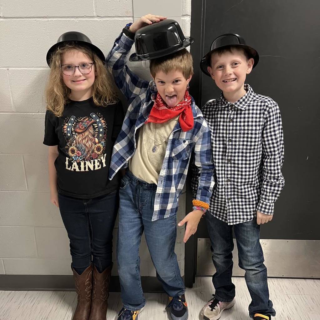 Three Unity third-graders posing playfully in their concert attire, featuring black bowler hats and western-style accessories like a red bandana and cowboy boots.
