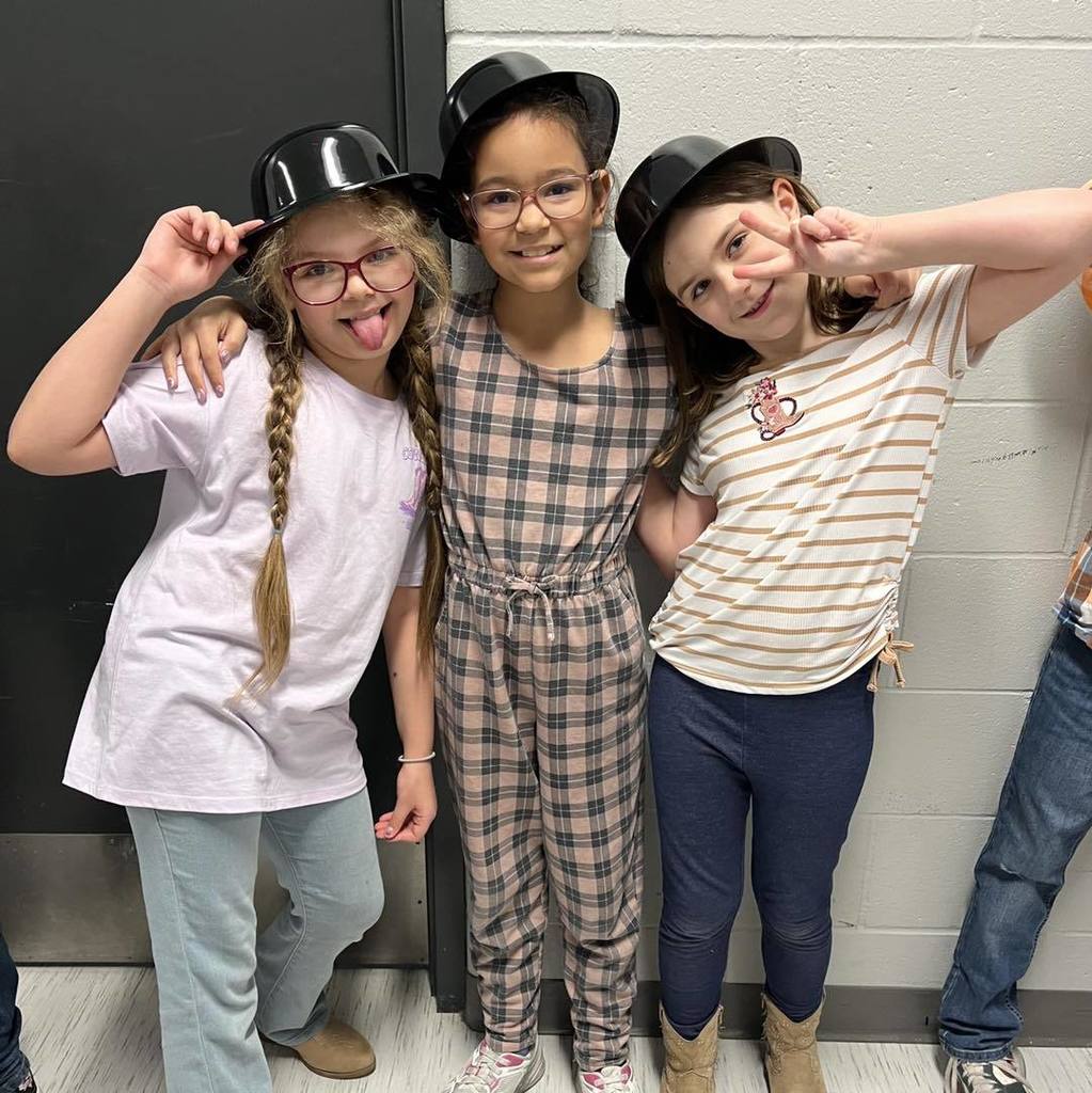 Three Unity third graders wearing black bowler hats and posing playfully for a photo in a school hallway.