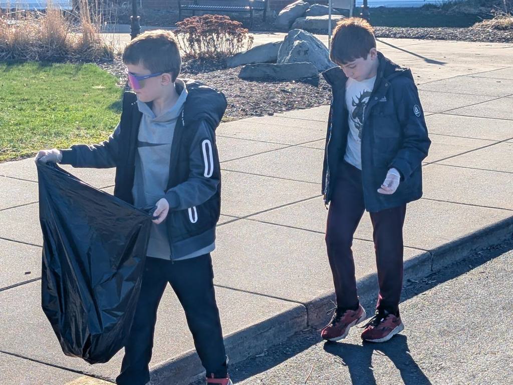 Two young students working together to transport a large bag of trash across the sunny Unity campus during the Student Council spring cleaning event.