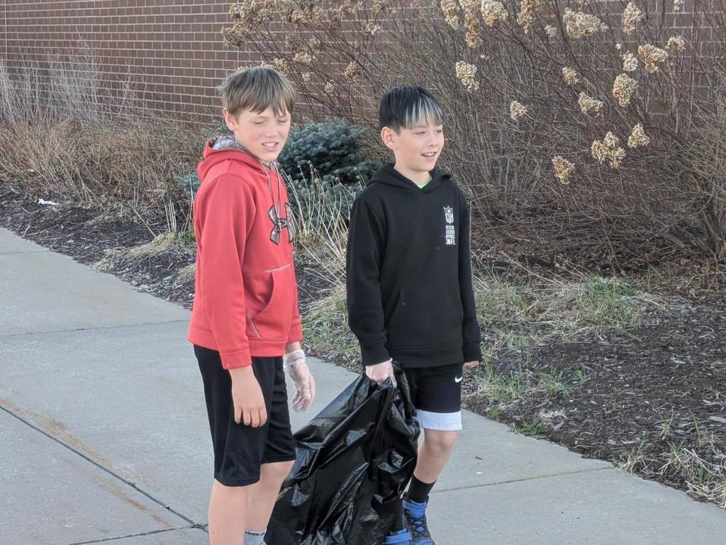 Two young students participating in the Student Council spring cleaning event, carrying a bag of trash across the school grounds.