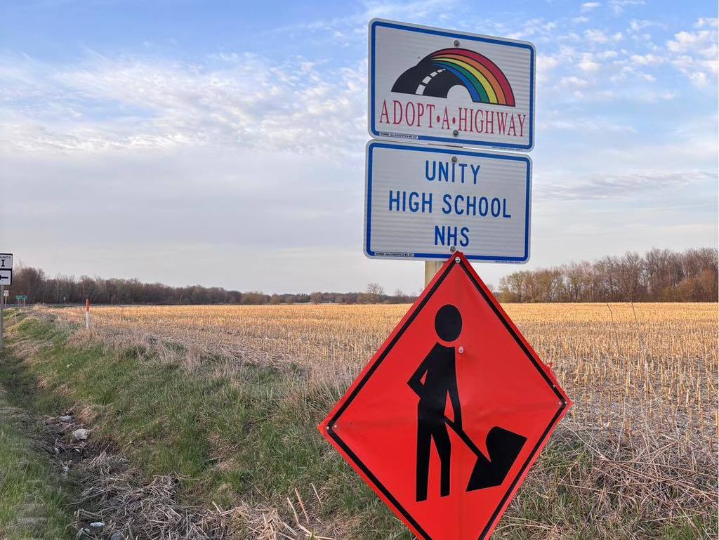 An Adopt-A-Highway sign for Unity High School NHS stands next to an orange "Men Working" diamond sign along a rural road during the Earth Day cleanup.