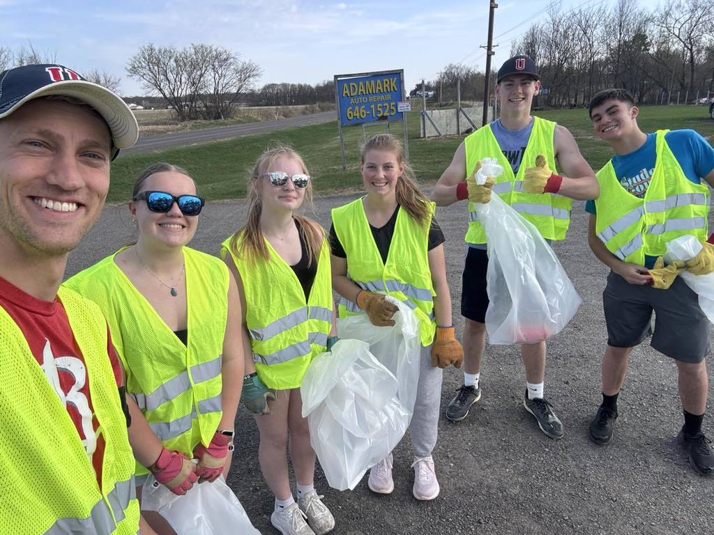 A group selfie of Mr. Mathias and five Unity NHS students posing together on a gravel roadside. They are all wearing neon yellow safety vests and work gloves, smiling as they hold up large clear bags filled with trash collected during their Earth Day highway cleanup.