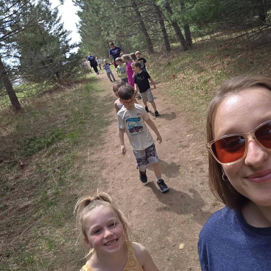 A teacher and a line of Unity Kindergarten students exploring a wooded trail on a sunny day during their Earth Day nature walk.