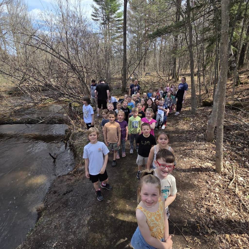 A class of Kindergarten students exploring the outdoors for Earth Day, lined up along a scenic forest path next to a stream to observe nature.