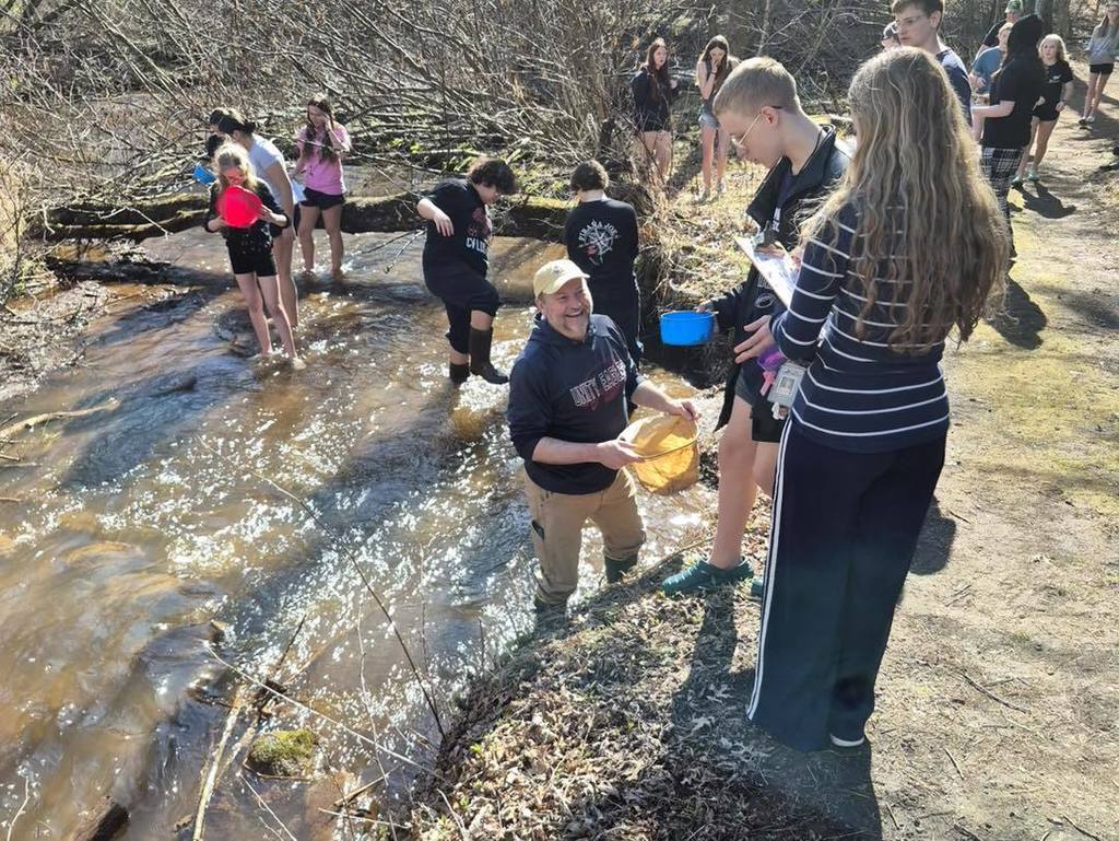 A Unity 7th grade student in a striped shirt and a teacher in a Unity Eagles shirt work together at Rice Creek. The teacher wades in the shallow water, handing a yellow collection net to the student, who stands on the sunlit bank ready to index their findings.