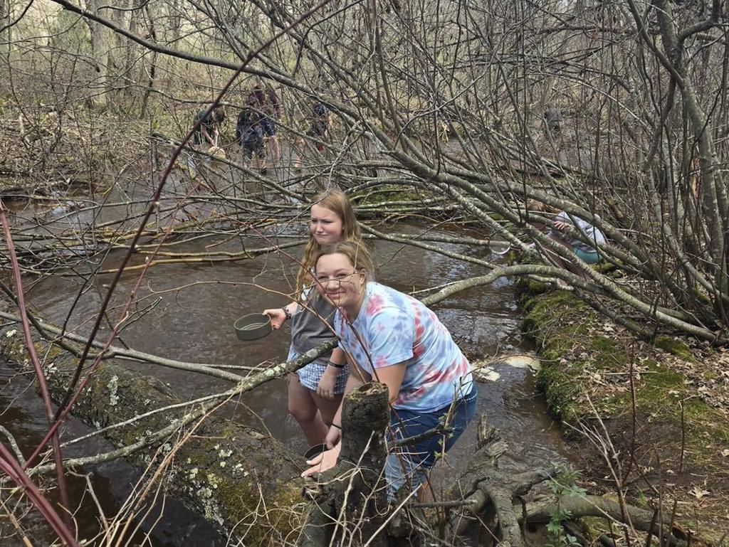 A close-up of two Unity 7th grade girls smiling while exploring a muddy section of Rice Creek. One student in a tie-dye shirt leans against a fallen log while the other holds a small collection container, with more classmates visible in the background investigating the stream.