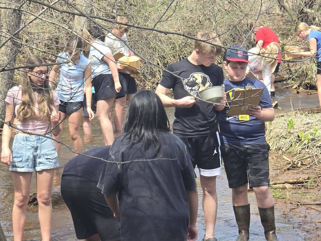 A group of Unity 7th grade students gathered in the shallow water of Rice Creek to examine their findings. Two boys in the center look closely at a clipboard and a scientific strainer, while other classmates in the background use nets to search the stream bed for macro-invertebrates.