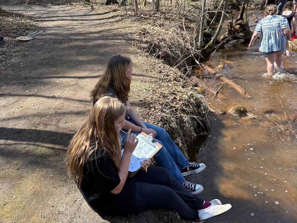Unity science students indexing their findings on clipboards while stationed along the edge of Rice Creek during an Earth Day stream survey.