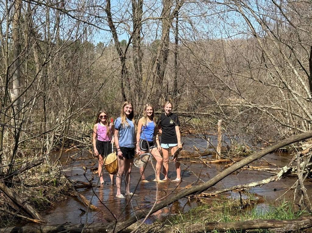 Four Unity 7th grade girls posing together while wading in Rice Creek. They are holding scientific collection nets and wearing water shoes, surrounded by fallen branches and the budding spring forest as they search for macro-invertebrates.