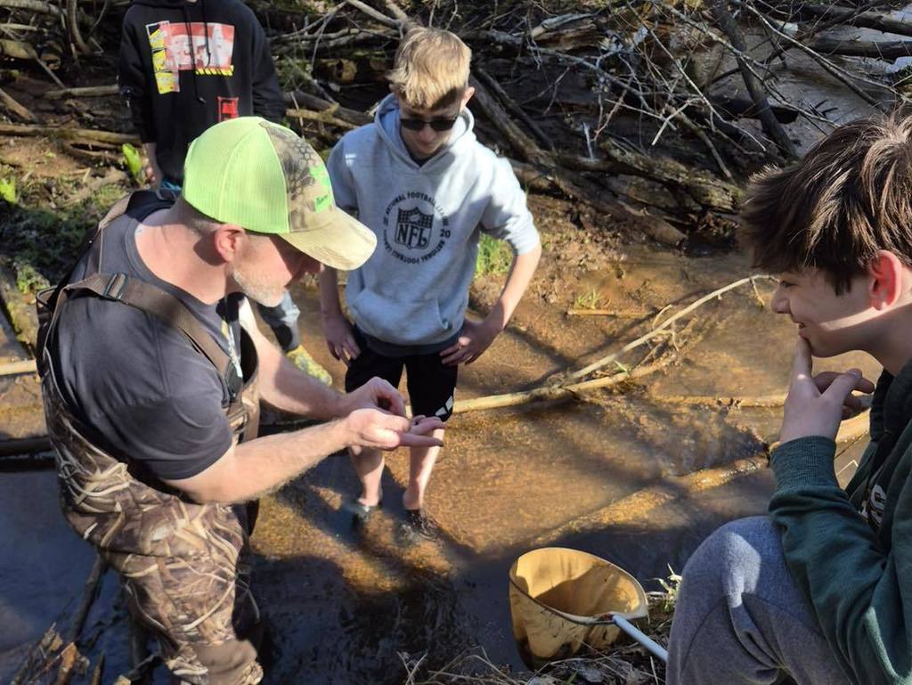 A close-up of a teacher in a neon green hat and chest waders showing a small aquatic find in his palm to two Unity 7th grade students. The students look on with curiosity as they stand in the shallow, clear water of Rice Creek during their Earth Day stream survey.