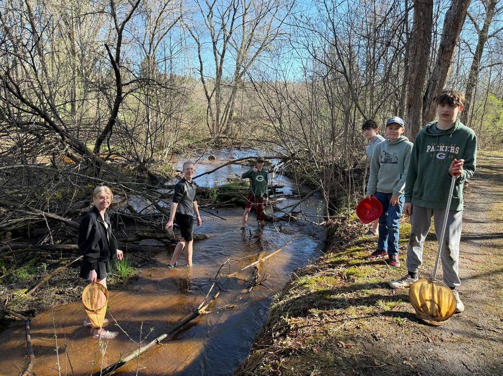 A group of Unity 7th grade students spread out at Rice Creek for a stream survey. Some students wade in the shallow, rocky water with collection nets, while others stand on the dirt path with buckets and clipboards, all working together to collect macro-invertebrate samples.