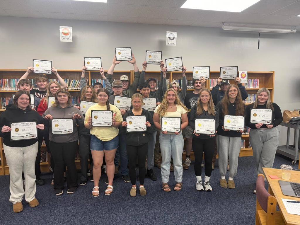 A large group of 21 Unity High School students standing together in the school library, smiling and proudly holding up their Youth Apprenticeship completion certificates. They are arranged in two rows in front of bookshelves, celebrating their success at the annual Youth Apprenticeship Banquet.