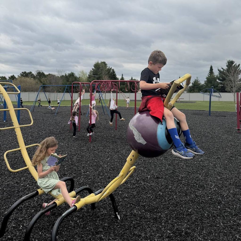 Two Unity students sitting on a yellow and purple spring-rider playground toy, deeply focused on reading their books during recess. In the background, other students play on the jungle gym and swings under a cloudy sky.
