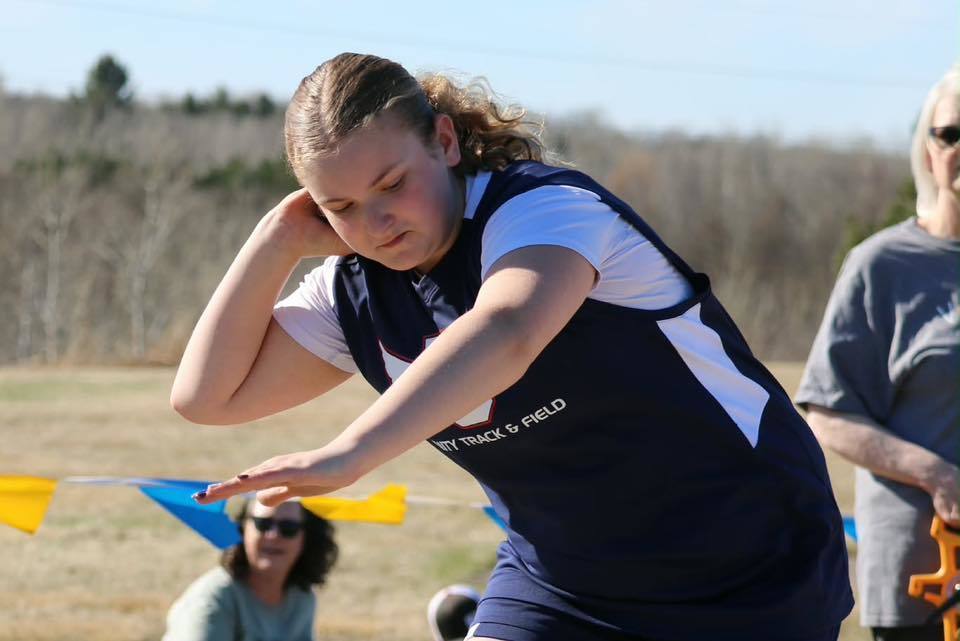 A Unity Middle School girl track athlete in a blue and white "Unity Track & Field" jersey preparing to throw a shot put. She is focused and holds the metal ball tucked by her neck, ready to launch, with a sunlit field and spectators in the background.