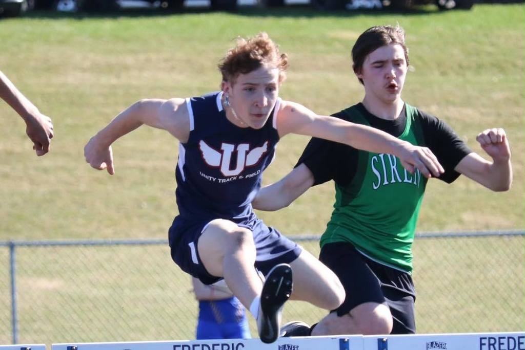 A Unity Middle School boy track athlete in mid-air as he leaps over a hurdle during a race. He is wearing a blue Unity Track and Field jersey and is focused on the finish line, with a competitor from Siren in a green jersey running closely behind him.