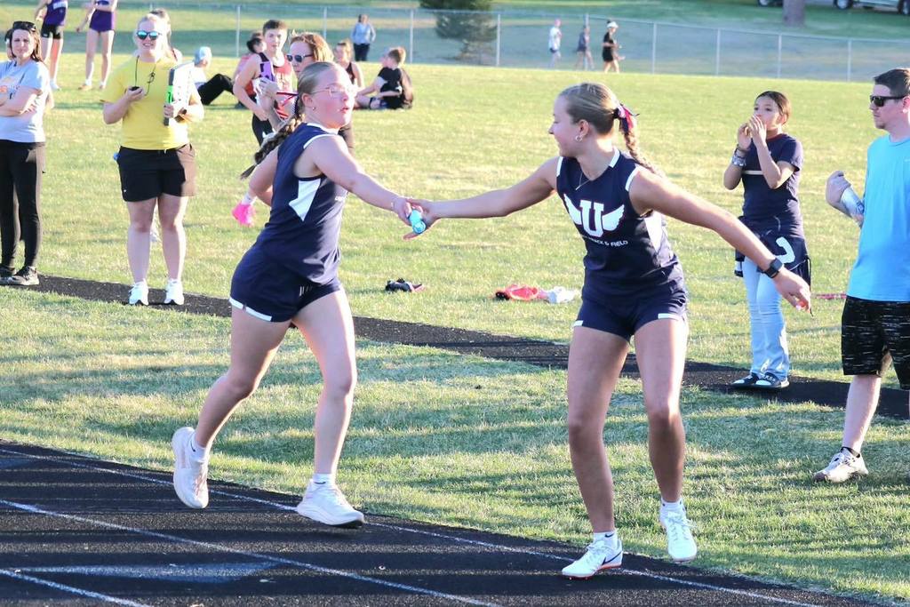 Two Unity Middle School girls' track athletes performing a relay baton handoff during a race. One student in a blue Unity uniform stretches her arm back to receive the light blue baton from her teammate as they run on the black track under the late afternoon sun.