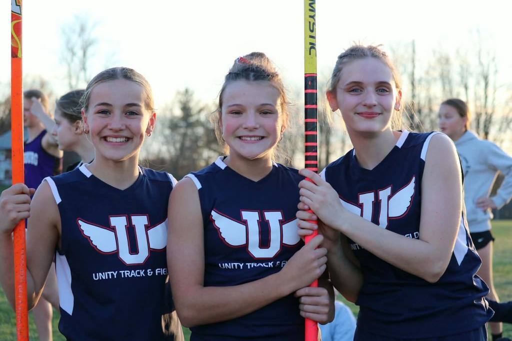 Three Unity Middle School girls' track athletes smiling together during an outdoor meet. Two of the students are holding onto tall, vertical poles, and all three are wearing blue and white "Unity Track & Field" jerseys. The background shows a sunlit field with other students and a line of budding trees.