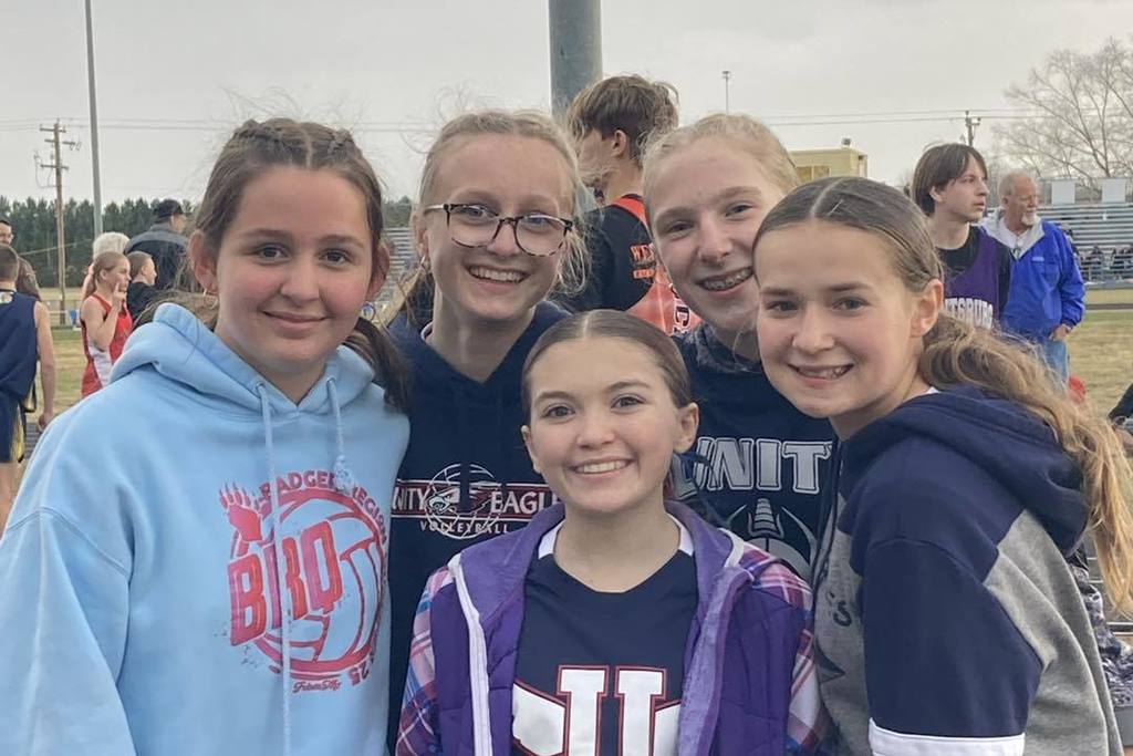 A group of five Unity Middle School girls' track athletes smiling together for a photo. They are dressed in a mix of blue Unity uniforms and warm-up gear, including a light blue "Badger" hoodie and a Unity Eagles sweatshirt, standing in front of an outdoor track with other students in the background.