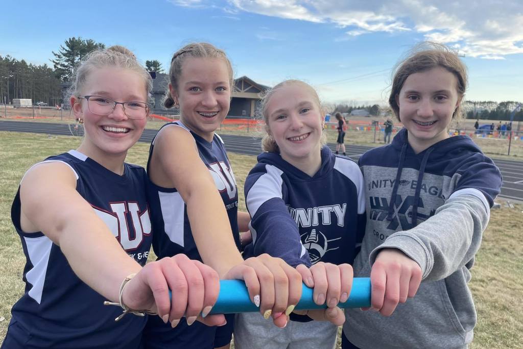 Four members of the Unity Middle School girls' track team smiling together on the track. They are all holding onto a single blue relay baton, symbolizing teamwork. Three students are in blue Unity uniforms and one is in a grey Unity Eagles sweatshirt.