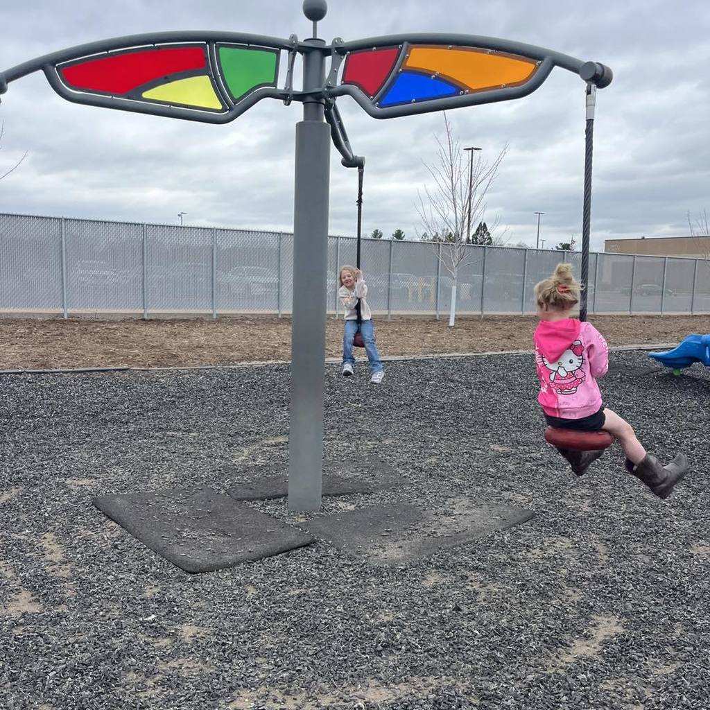 For the fourth photo in your 1st grade series, showing the students on the colorful spinning swing, here are your alt text options for Thrillshare:  Option 1: Detailed & Descriptive (Recommended) Two Unity 1st grade students enjoy a ride on a modern playground swing. One student in a pink Hello Kitty hoodie sits on a red seat in the foreground, while another classmate swings in the background. The equipment features unique stained-glass style panels in red, yellow, green, and blue that catch the light.