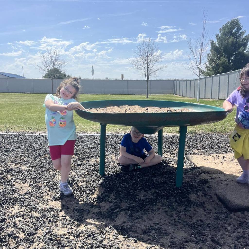 Three Unity 1st grade students enjoying a sunny day on the playground. One girl in a light blue shirt smiles while leaning on a large, circular green sand table, while a boy sits in the shade underneath it. Another student in a purple shirt stands on the opposite side, all soaking up the spring sunshine.