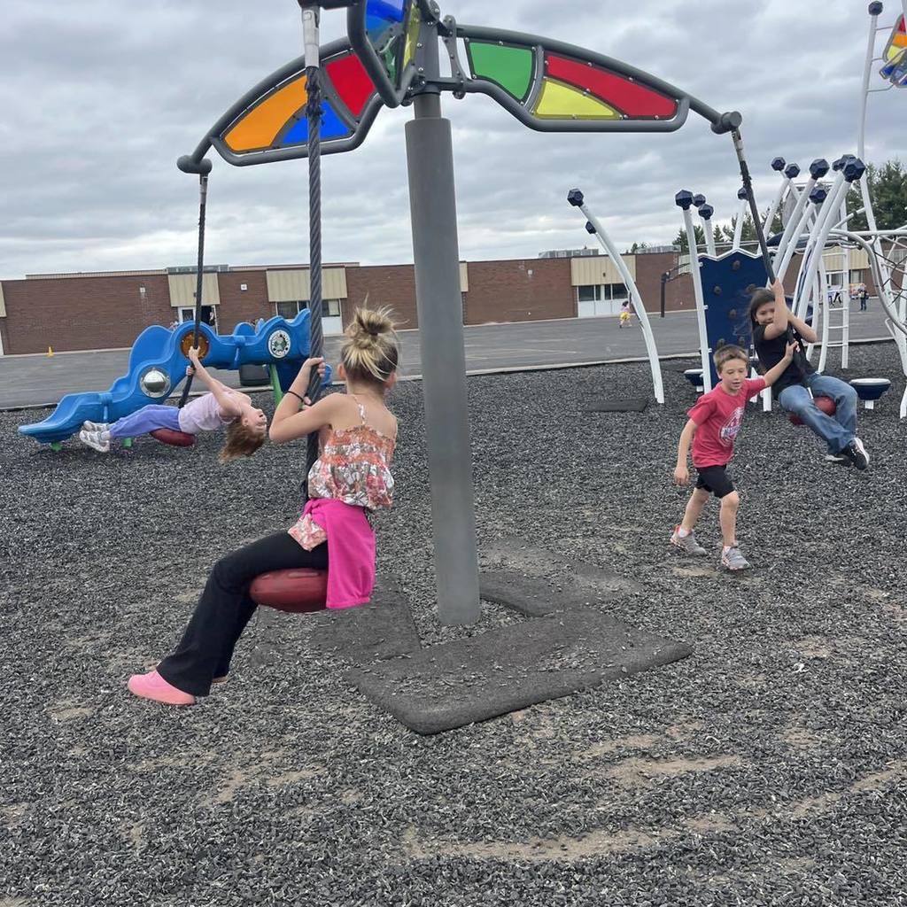 A group of Unity 1st grade students playing on a large, modern spinning playground structure. One girl in a floral shirt and pink leggings sits on a red seat in the foreground, while three other students hang from the ropes and sit on the rotating arms. The spinner features colorful stained-glass style panels at the top against a cloudy sky.