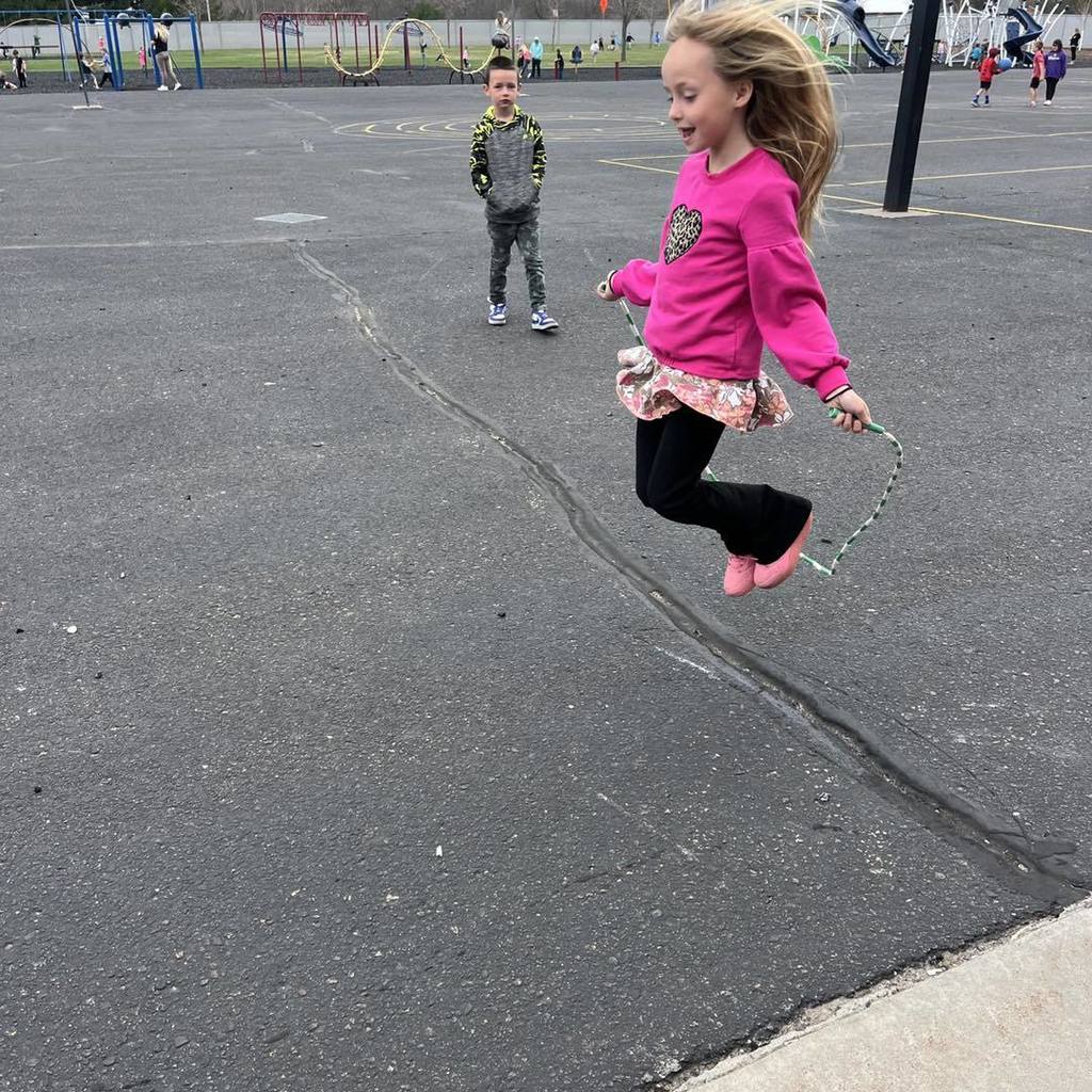 A Unity 1st grade girl in a bright pink shirt and heart-patterned skirt caught in mid-air while jumping rope on the school playground. A boy in a camouflage sweatshirt stands nearby watching, while other classmates play in the background on a sunny day.
