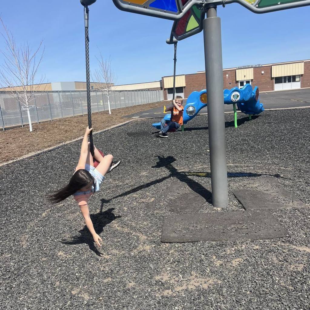 A Unity 1st grade girl glides through the air on a rope zipline swing at the school playground. Another student is visible in the background on a second swing, both enjoying the sunshine and outdoor activity on a clear spring day with the school building behind them.
