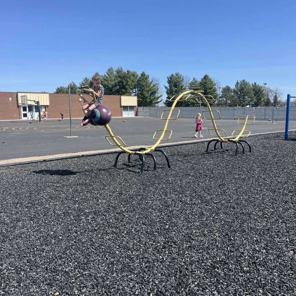 A Unity 1st grade student sits on a purple ball-shaped seat attached to a long, wavy yellow metal climbing structure on the playground. Another student walks in the background on the black rubber mulch, all enjoying a bright, cloudless spring day at the eleme