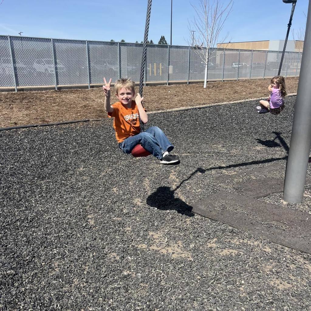A Unity 1st grade boy wearing an orange shirt and jeans flashes a peace sign while sitting on a red disc swing. He is soaring through the air on the playground under a bright blue sky, with a classmate on a second swing and the school building in the background.