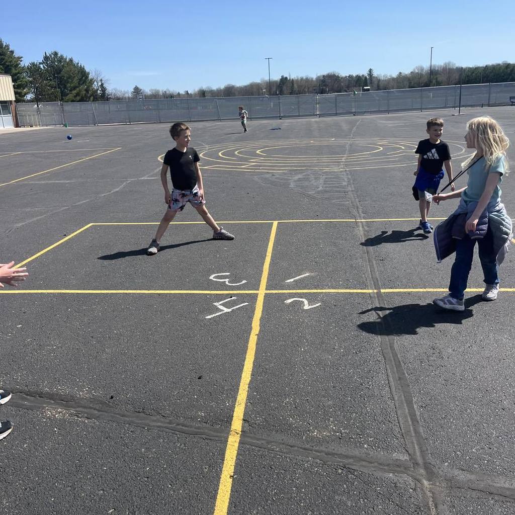 Three Unity 1st grade students playing a game of four square on a sunny asphalt playground. Two boys and one girl are positioned in their numbered squares, enjoying the warm spring weather under a bright blue sky with the school building in the background.