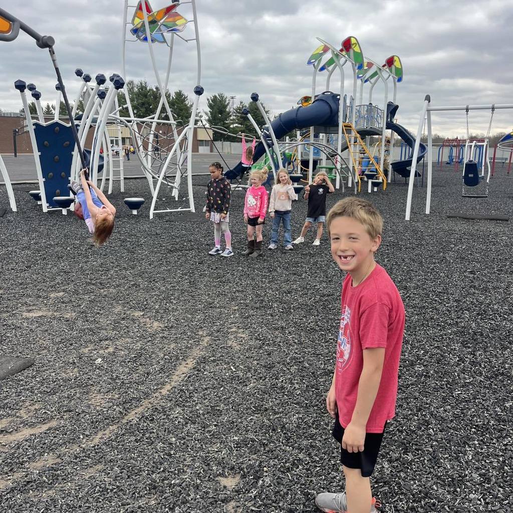 A smiling Unity 1st grade boy in a red t-shirt stands in the foreground of a busy playground. Behind him, a girl in a blue shirt hangs upside down on a zipline swing, and a group of classmates stand near a large blue climbing structure under a cloudy spring sky.