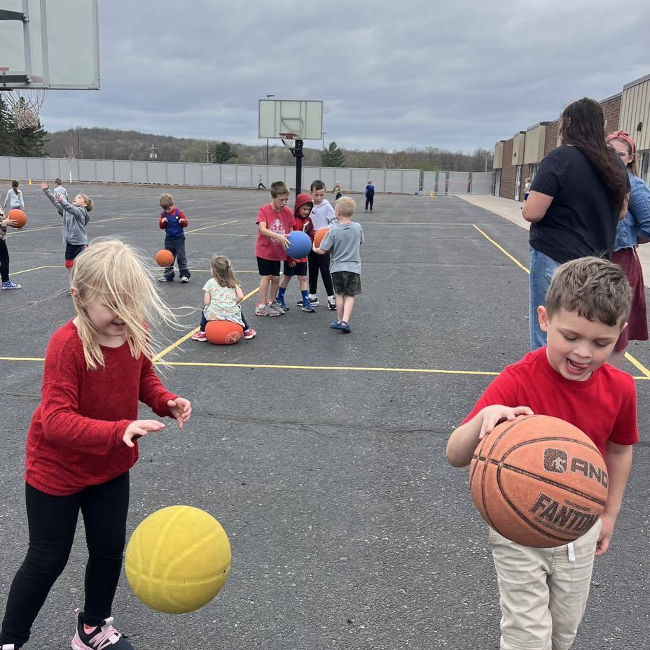 Unity 1st grade students playing with basketballs and playground balls on the school blacktop. In the foreground, a girl in a red sweater watches a yellow ball bounce while a boy in a red shirt holds a basketball. Other classmates and a basketball hoop are visible in the background under an overcast sky.