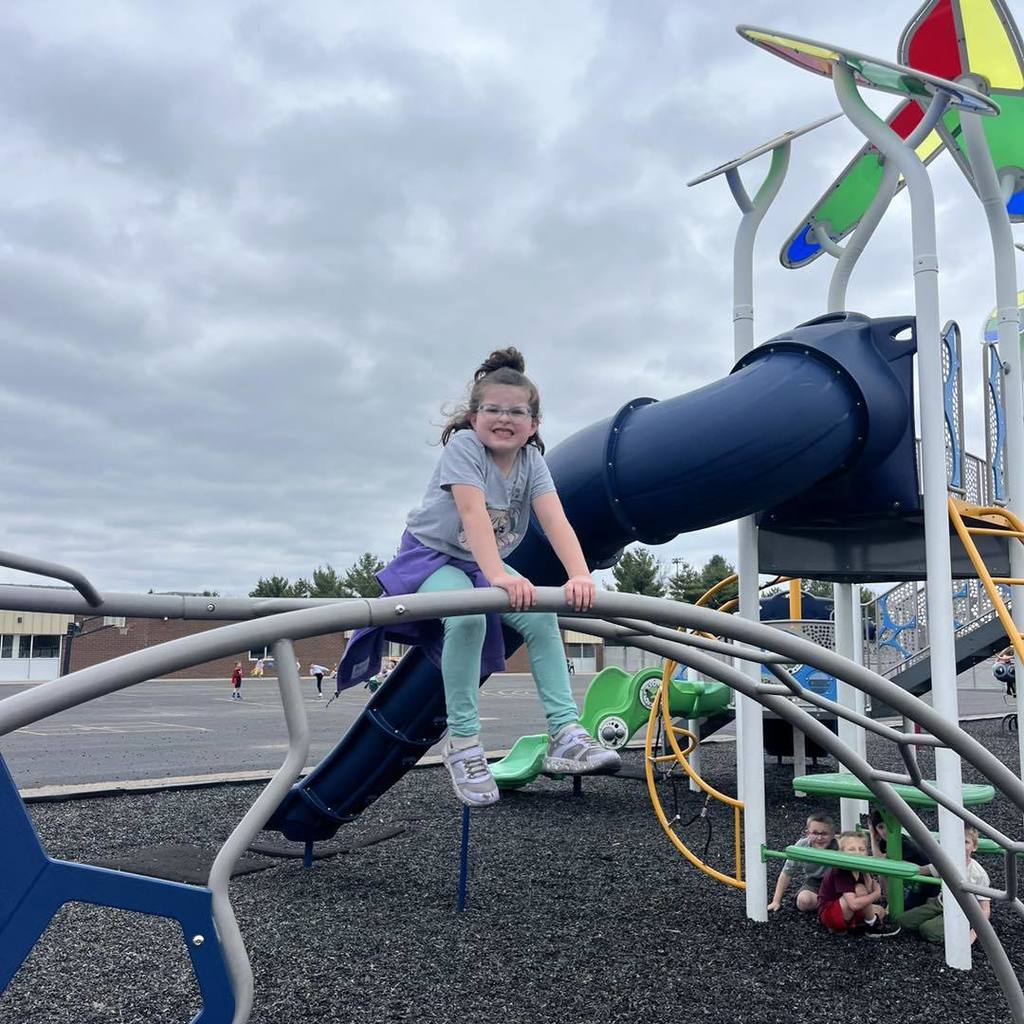 A Unity 1st grade girl with glasses and a purple jacket smiling as she climbs on top of a gray metal arched climbing structure. In the background, a large blue tunnel slide and a colorful playground set stand under a cloudy sky, with other students playing nearby on the black rubber mulch.
