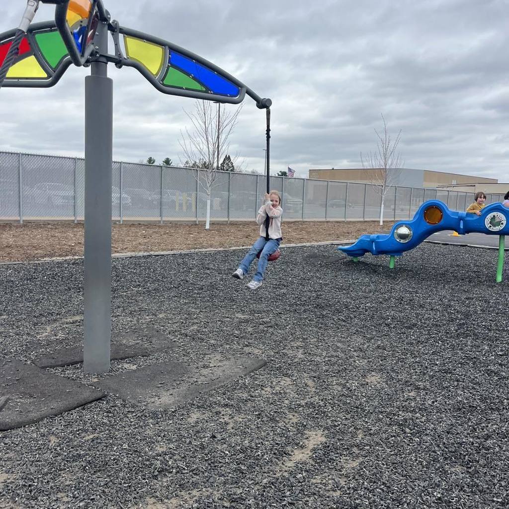 A Unity 1st Grade girl swinging on a rope zipline swing. She is smiling and holding onto the rope under a cloudy sky, with a colorful stained-glass style playground structure and the school building in the background.