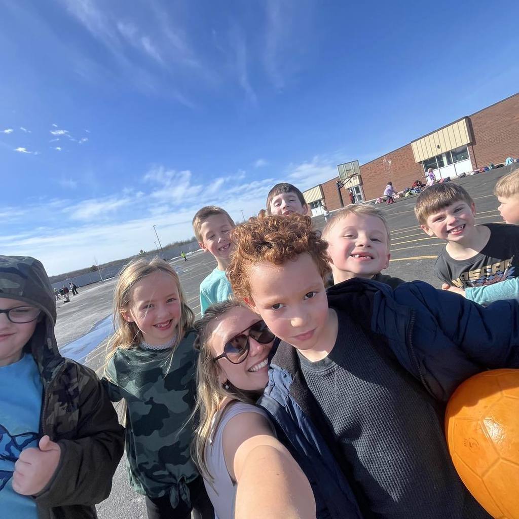 A joyful group selfie of a Unity staff member and several 1st grade students huddled together on the sunny playground. The students are smiling and making thumbs-up gestures under a bright blue sky, with the brick school building and the blacktop in the background.