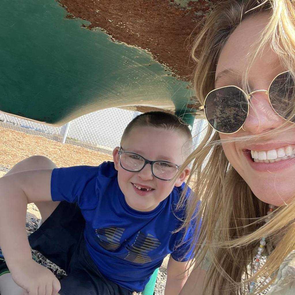 A close-up selfie of a smiling Unity staff member and a 1st grade boy on the playground. The staff member is wearing gold-rimmed sunglasses, and the student is wearing glasses and a blue t-shirt. They are posing together in the sunshine under the shade of a green playground structure.