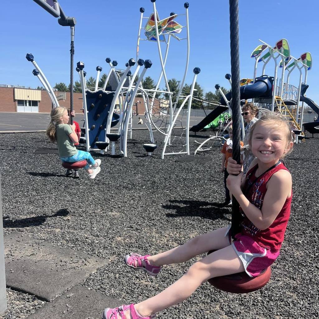 A Unity student smiling while riding a zipline swing during a sunny outdoor recess. The photo captures the excitement of 1st graders enjoying the spring weather on the school playground.