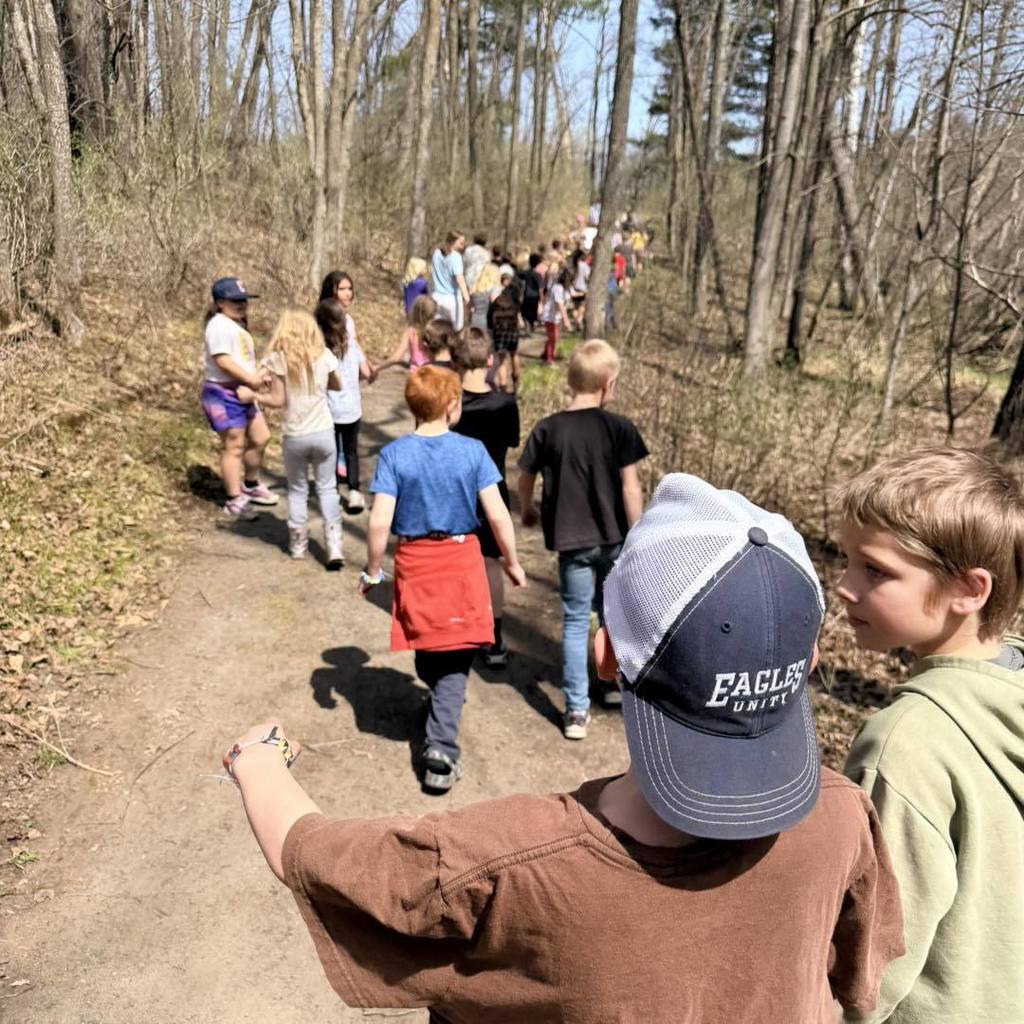 A line of Unity 2nd grade students hiking down a dirt nature trail through a sunlit forest. A boy in a brown shirt and "Eagles" hat leads the group in the foreground, as the line of classmates stretches into the distance between tall, budding trees.