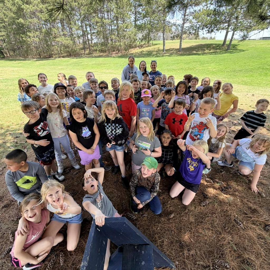 A large group of Unity 2nd grade students and their teachers posing for a photo under tall pine trees. The students are gathered on a bed of pine needles, smiling and making silly faces for the camera during their Earth Day nature hike on a sunny day.