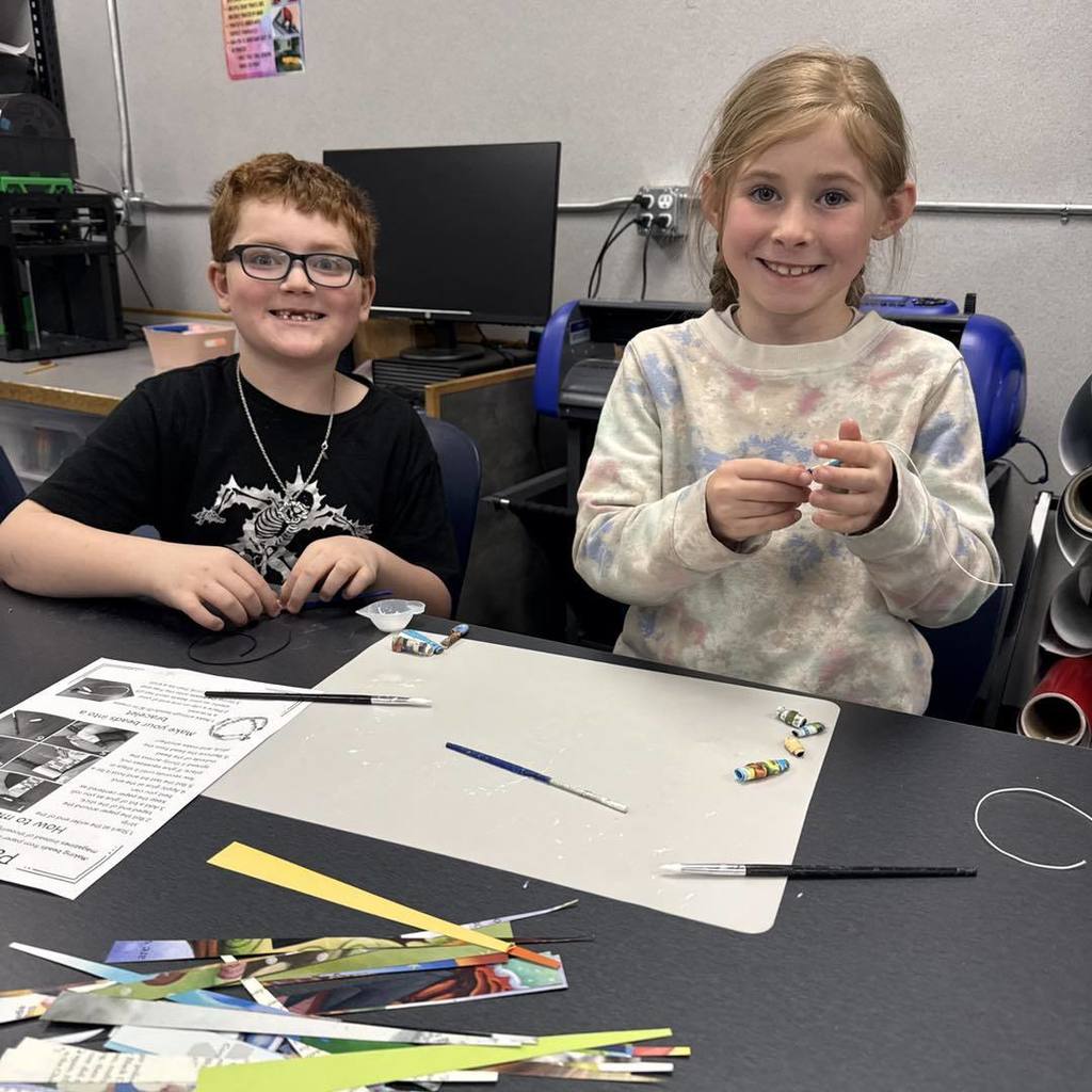 Two Unity 2nd grade students, a boy in a black t-shirt and a girl in a colorful patterned sweatshirt, smile while making Earth Day bracelets. They are sitting at a black desk covered in colorful paper strips, beads, and an instruction sheet titled "Make your own paper bead."