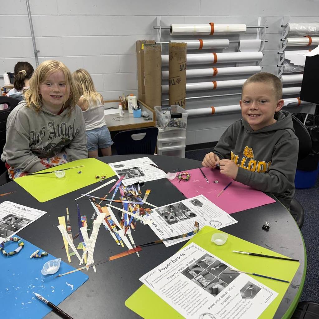 Two Unity 2nd grade students—a girl in a grey "Eagles" sweatshirt and a boy in a grey hoodie—smile at a table while making Earth Day bracelets. The table is covered in colorful paper strips and instruction sheets for making paper beads. In the background, large rolls of paper are mounted on the wall.